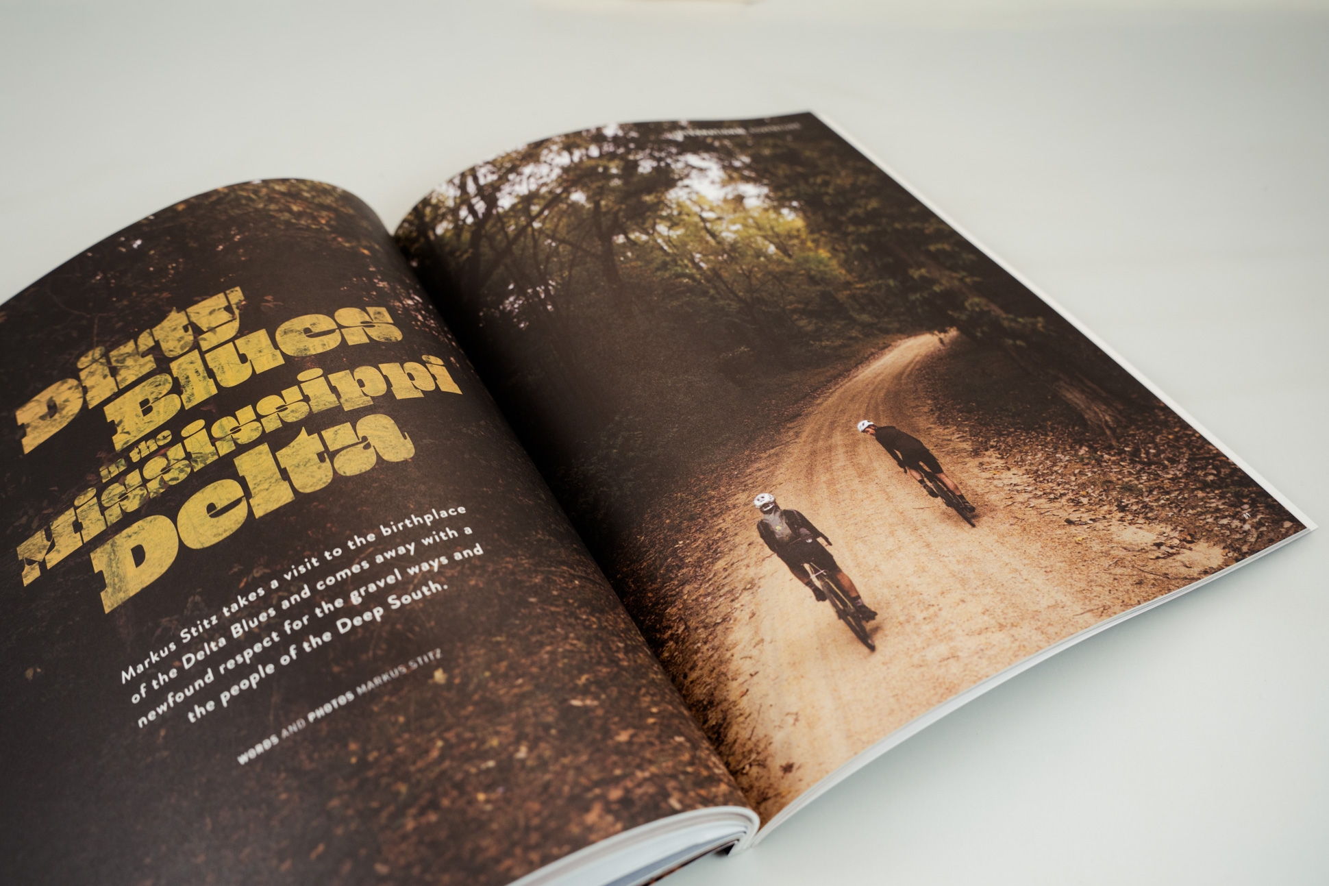 A close-up of an open magazine page featuring the title 'Dirty Blues in the Mississippi Delta' with two cyclists riding on a dirt path through a wooded area.