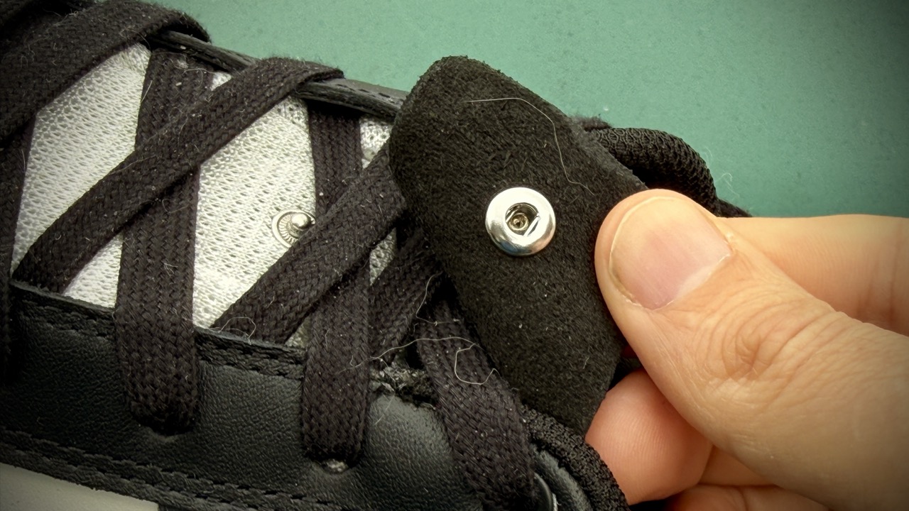 Close-up of a hand adjusting a black lace tag on a sneaker, with dark laces and grey mesh material visible. Drift ARC