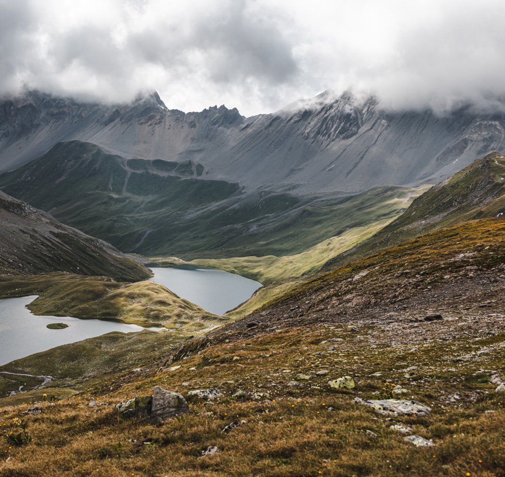 A small alpine lake set in green meadows beneath cloudy mountain peaks.