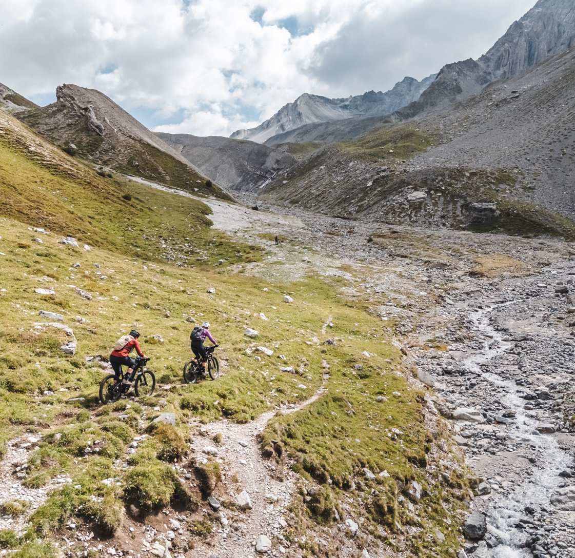 Two riders descending a rough stony trail in a wide glacial valley.