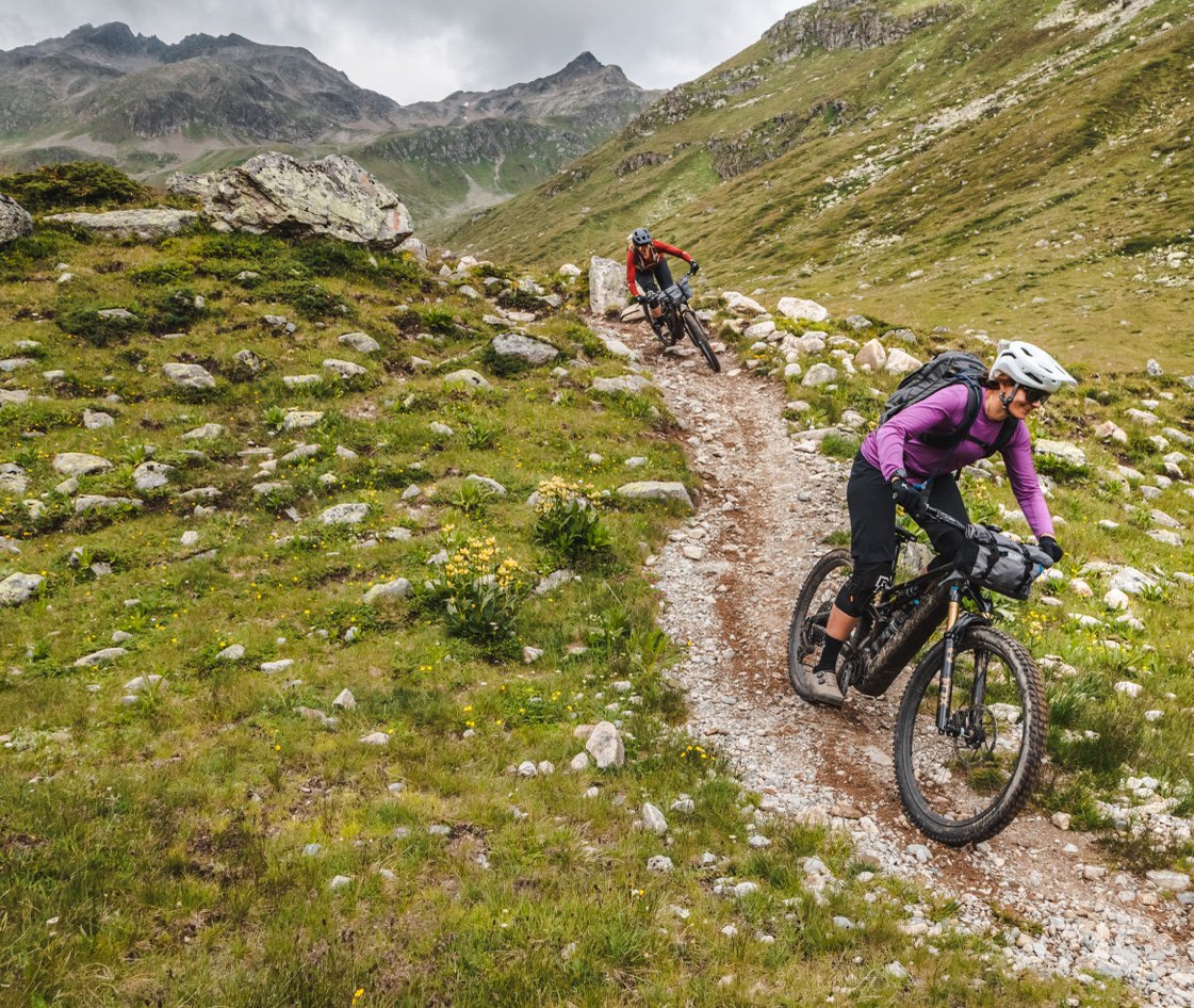 A rider in purple descending a worn singletrack across an open green mountainside.