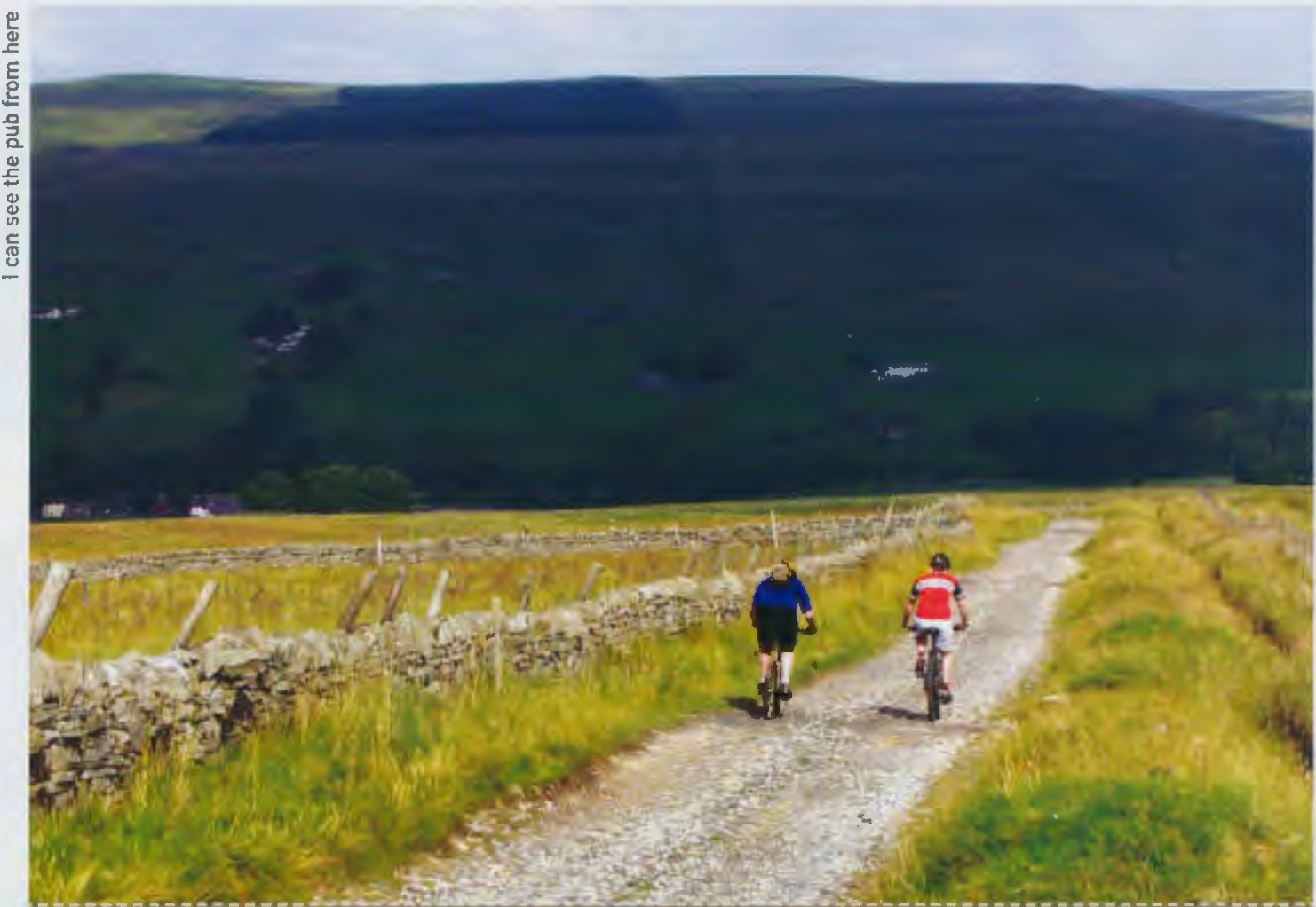 Two cyclists riding along a gravel path in a rural landscape, surrounded by green fields and stone walls.
