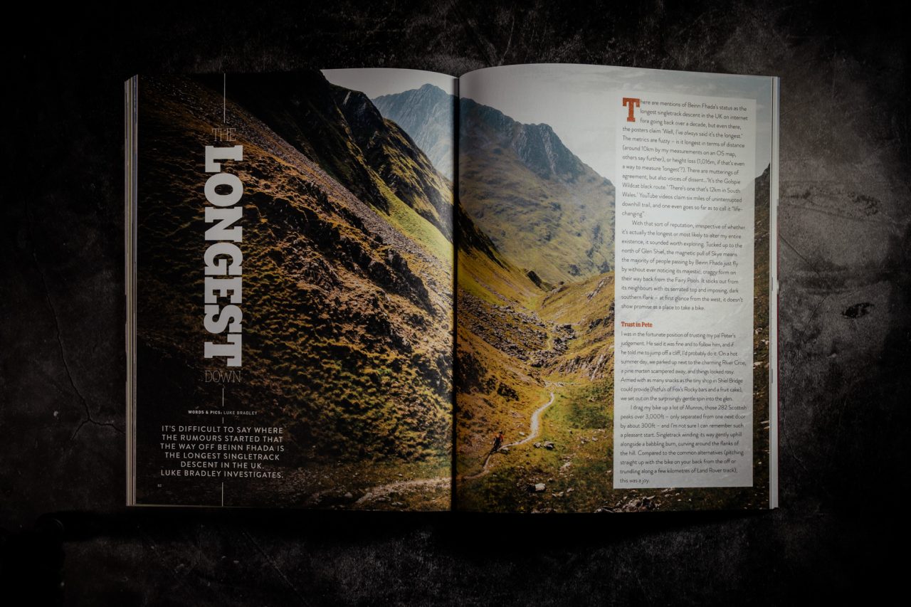 Double-page spread of 'The Longest' from Singletrack Magazine Issue 167, showing a lone mountain biker in an orange jacket on singletrack winding down a remote mountain valley