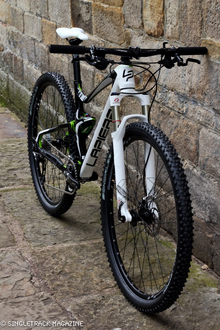 A modern mountain bike positioned against a stone wall, featuring a black and white frame with green accents, thick rubber tyres, and advanced suspension components.