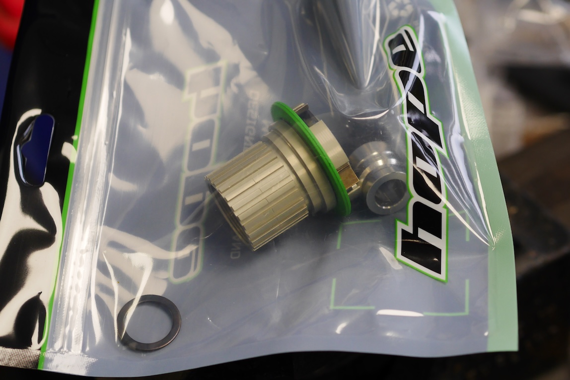 A close-up of a metallic component with a green gasket, placed inside a transparent plastic packaging with branding.