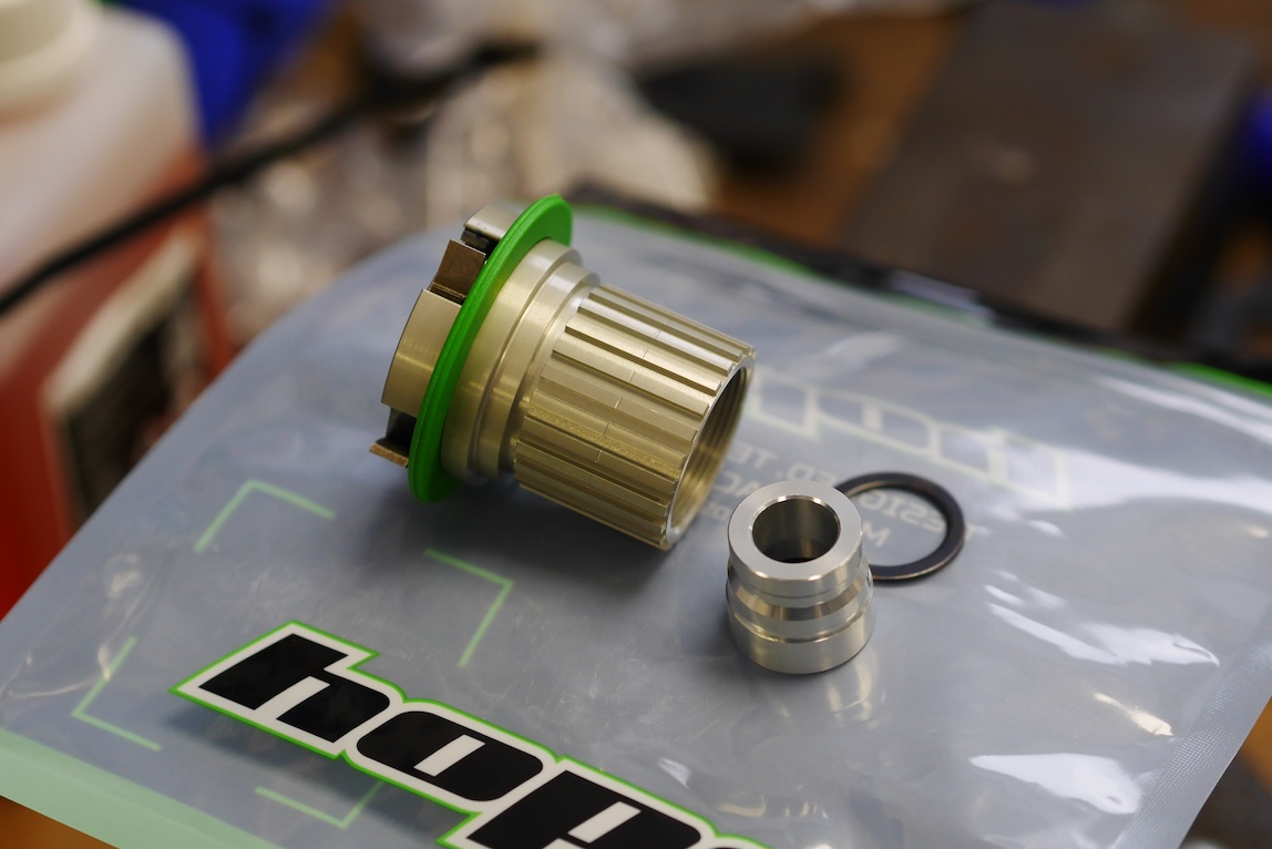 A close-up of bicycle components featuring a green-sealed aluminium hub and a silver spacer, placed on a branded plastic bag.