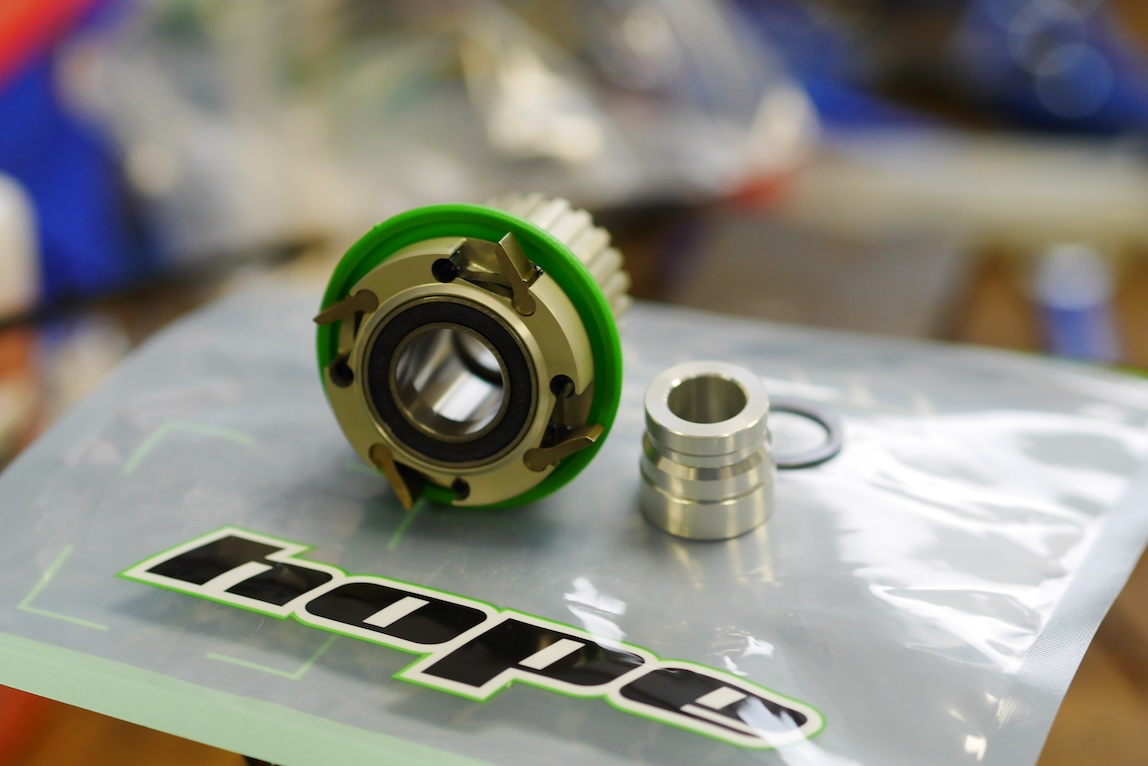 A bicycle component kit featuring a green and silver part, a silver spacer, and a black O-ring, displayed on a branded plastic bag.
