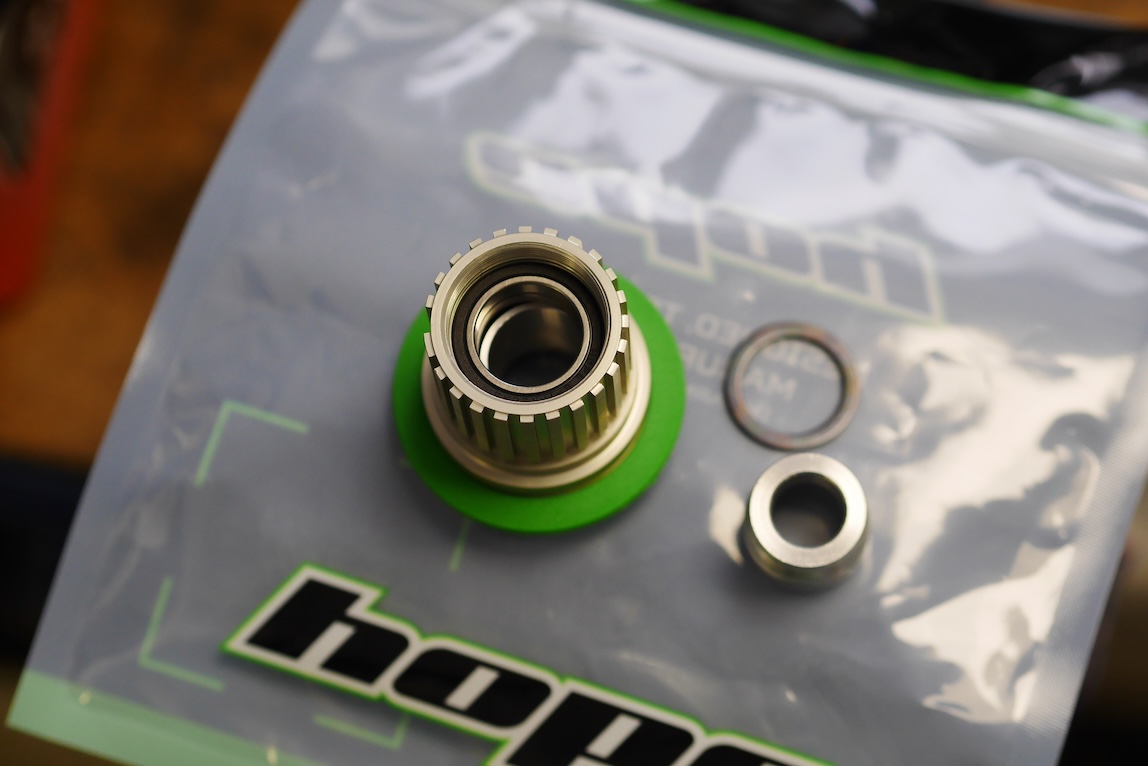 Close-up of mechanical components, including a metal part with grooves, a green rubber seal, and two small washers on a branded plastic bag.