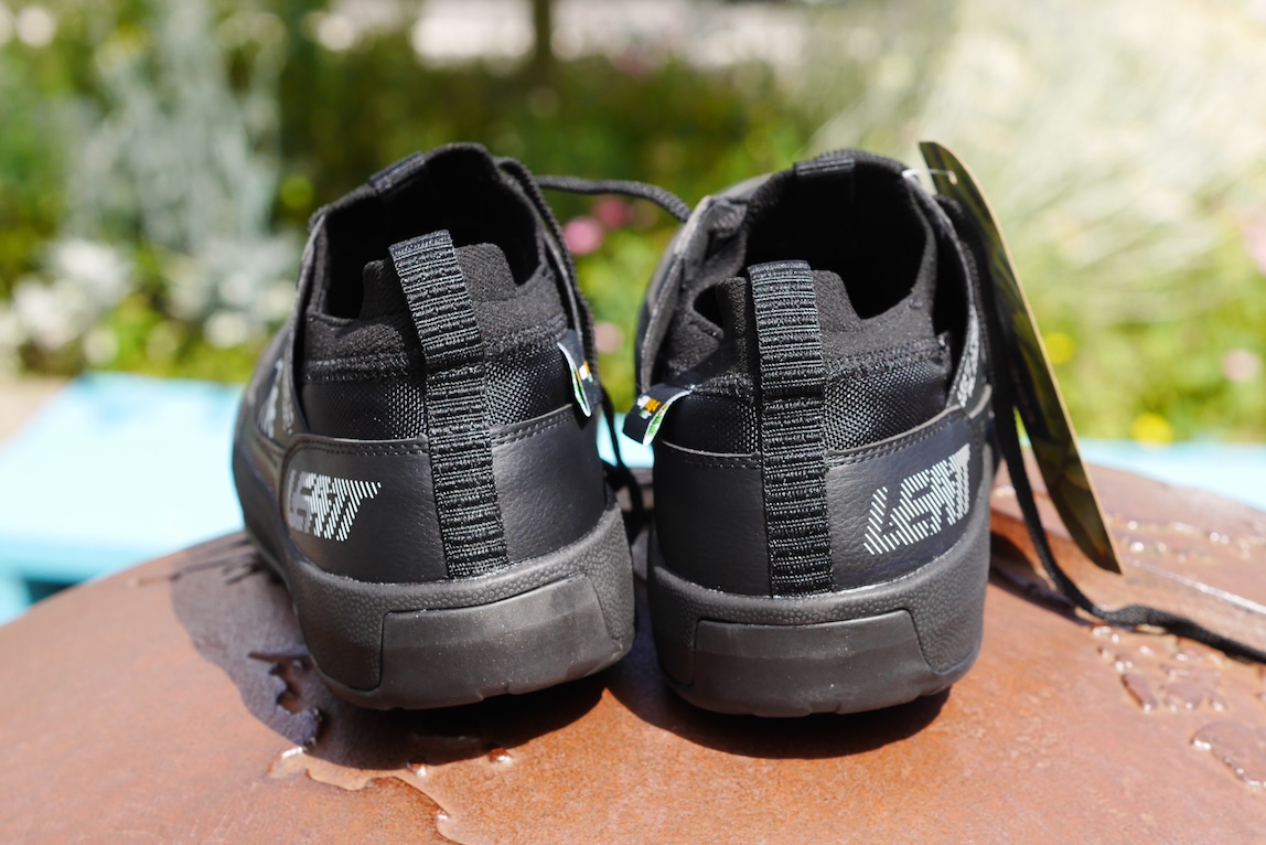 Back view of black sports shoes with reflective branding on the heel, set against a blurred garden background.