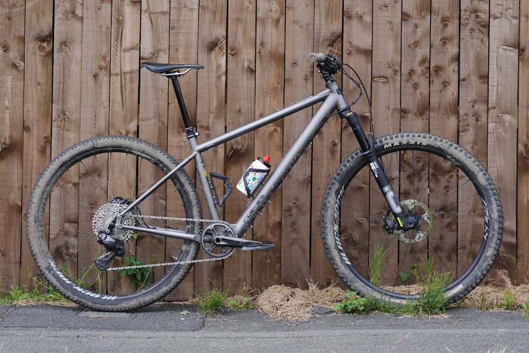 A mountain bike leaning against a wooden fence, featuring a grey frame, large tyres, and a water bottle mounted on the frame.