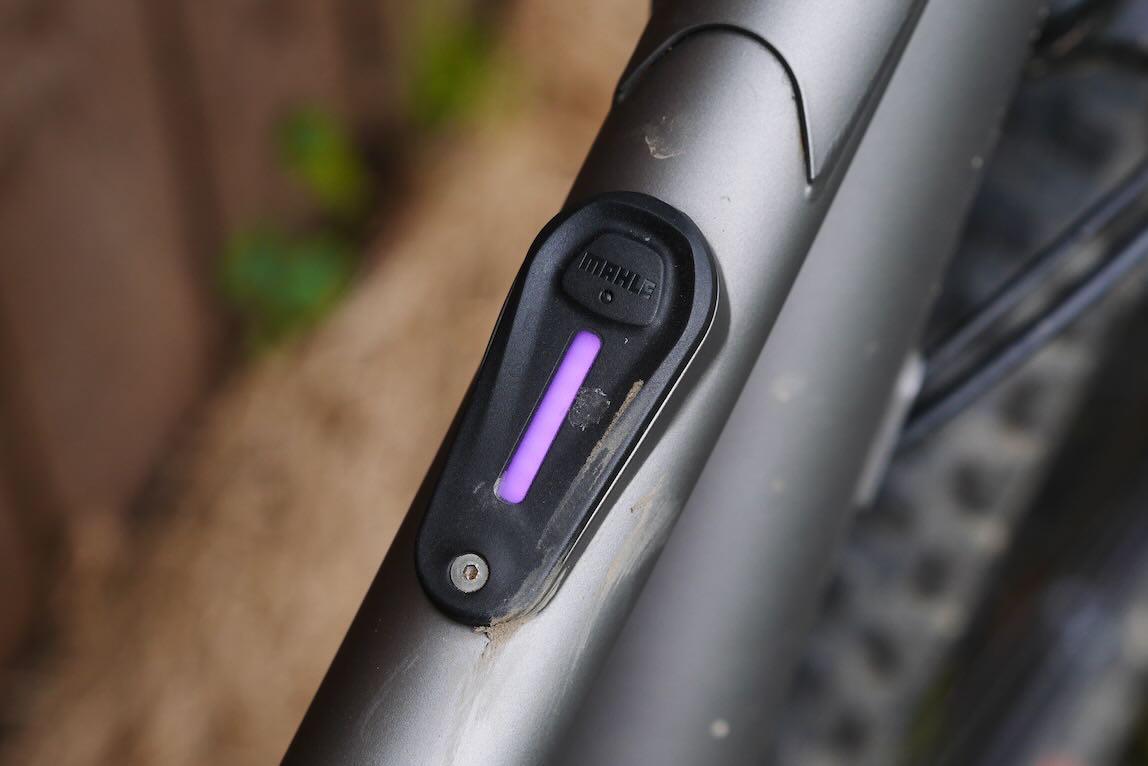 Close-up of a bicycle frame with a black component featuring a purple indicator, set against a blurred background.