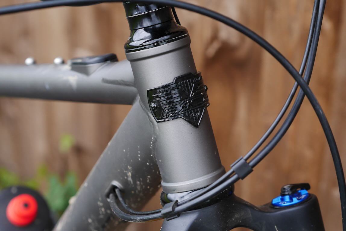 Close-up of a bicycle's head tube with a black emblem, showcasing the frame's detailing and cable routing.