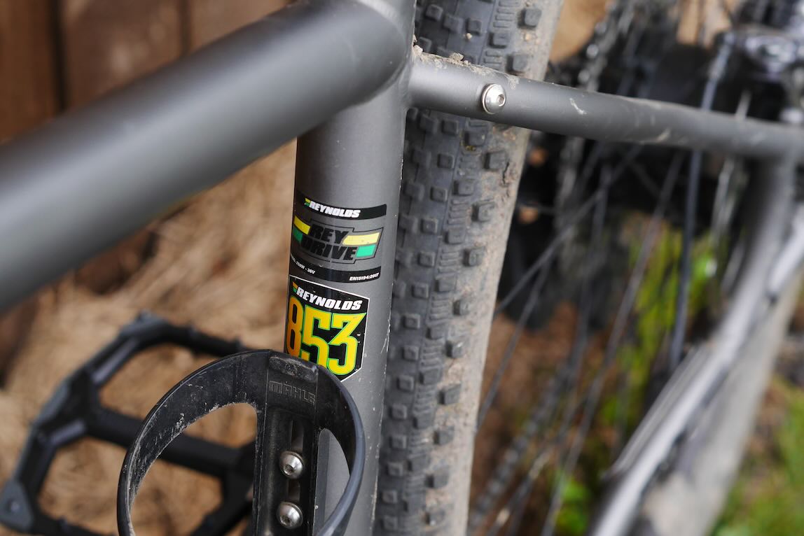 Close-up view of a mountain bike frame showing the Reynolds 853 sticker, a pedal, and a textured tyre in the background.