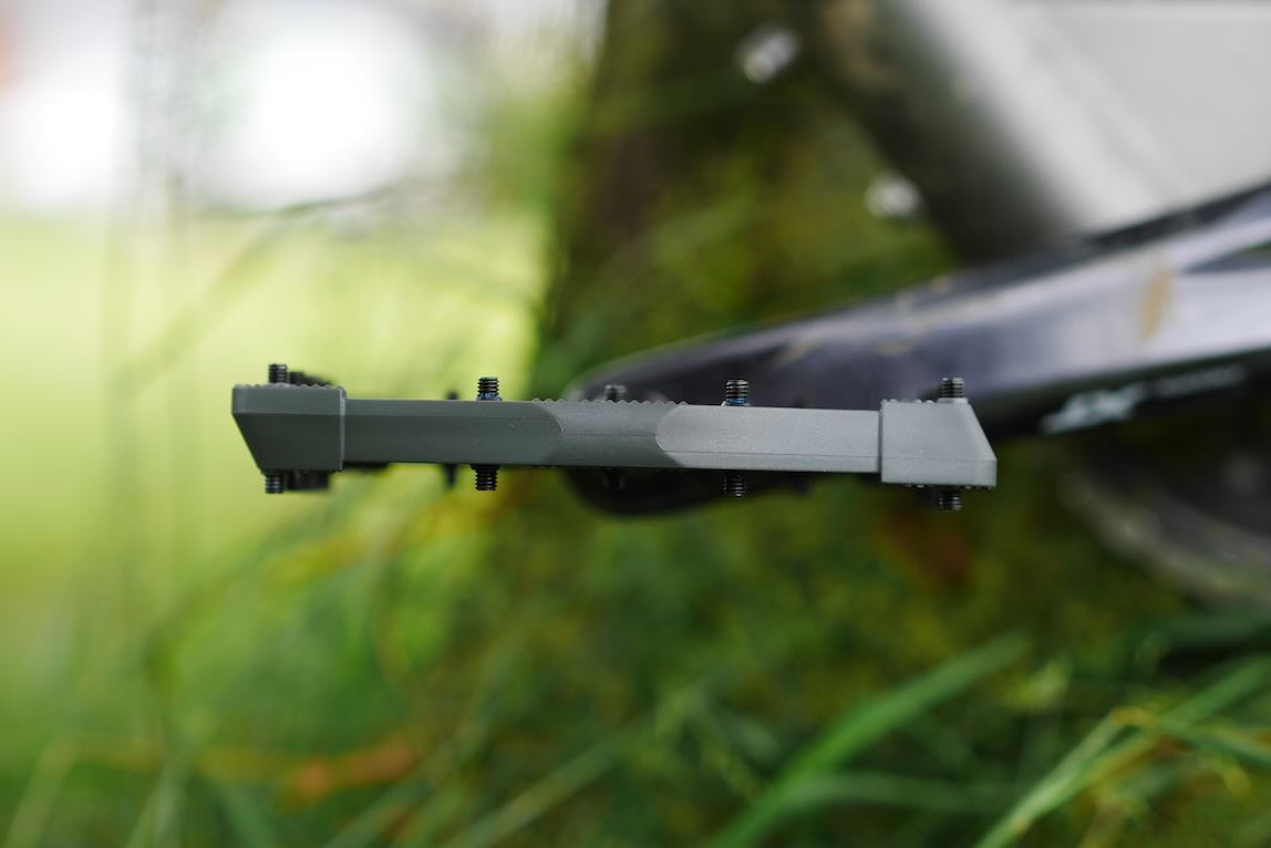 Close-up of a grey component, featuring multiple bolts, set against a green background with grass.