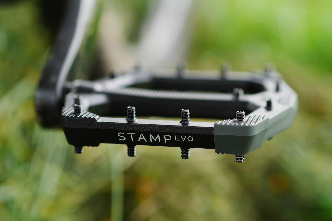 Close-up of a black and grey bicycle pedal with the text 'STAMP EVO' visible, resting on a grassy surface.