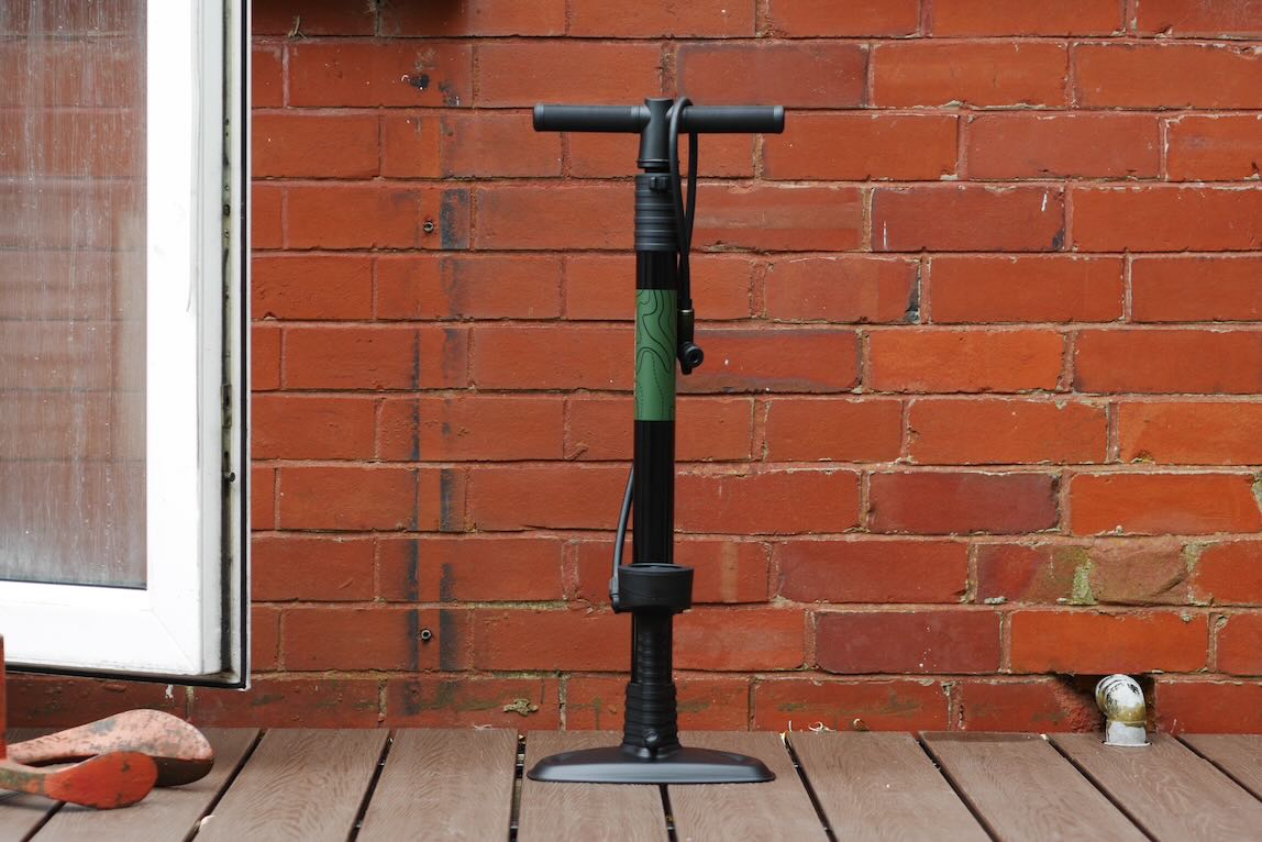A black and green pump standing against a red brick wall, with a wooden deck beneath it and an orange rubber object nearby.