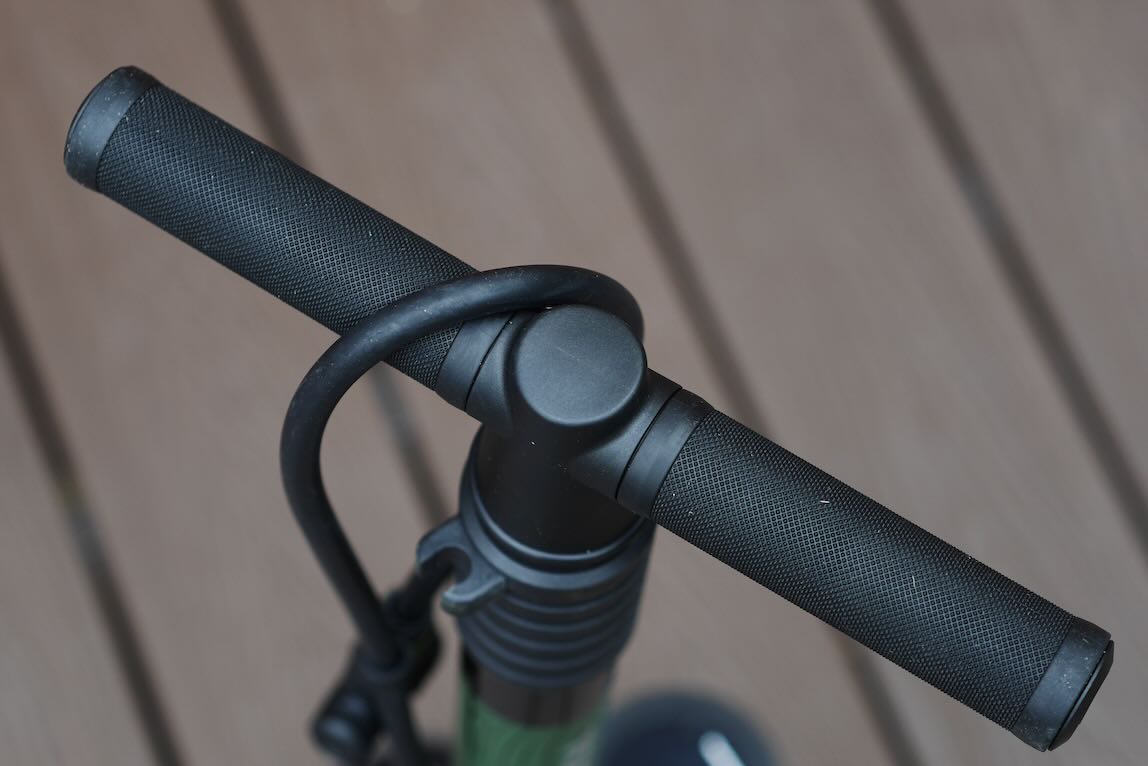 Close-up view of a black bicycle pump handle featuring textured grips, mounted on a green pump body, with a wooden surface in the background.