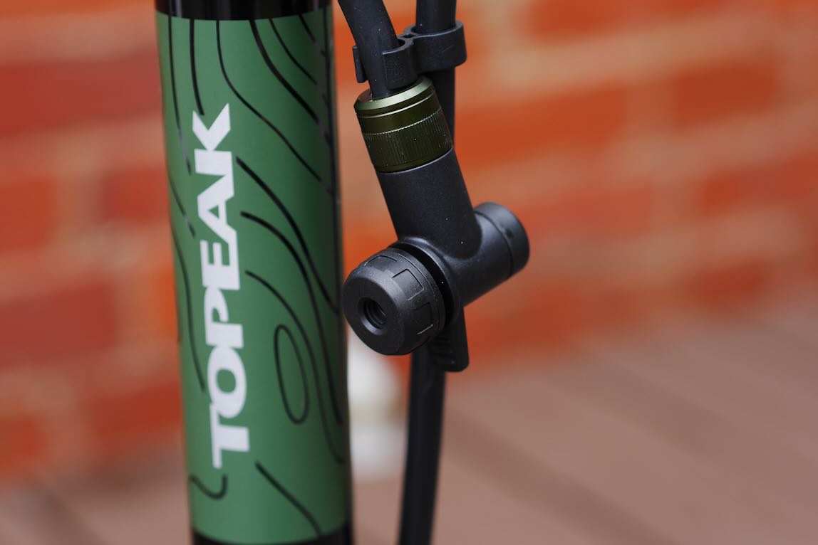 Close-up of a Topeak bike pump featuring a black nozzle and a green patterned pump body against a blurred brick background.