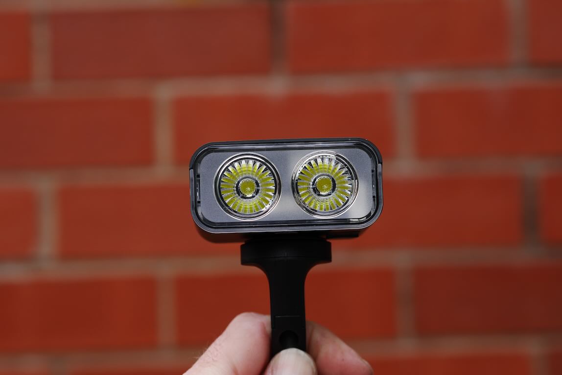 A hand holding a compact light with two circular LED bulbs in front of a textured red brick wall.
