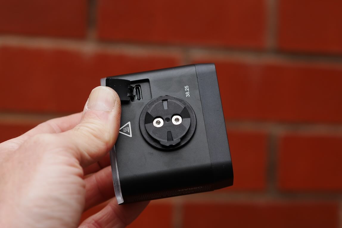 A person holding a compact black device with a circular connector and a small port, against a red brick background.