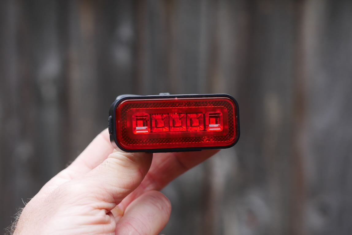A person holding a red bike rear light with four LEDs, against a blurred wooden background.