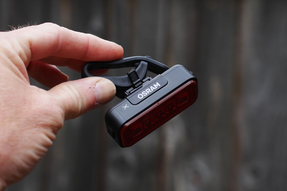 A hand holding a small black and red OSRAM bike light with a clip attachment against a blurred wooden background.