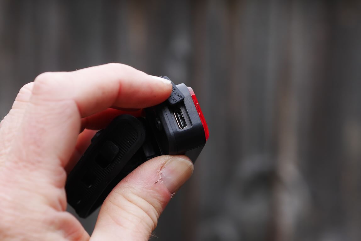 A close-up image of a person's hand holding a small electronic device, showing a USB port and a red element on the side, with a blurred wooden background.