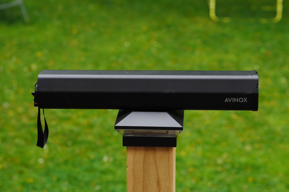 A black AVINOX device mounted on a wooden post, with a grassy background dotted with yellow flowers.