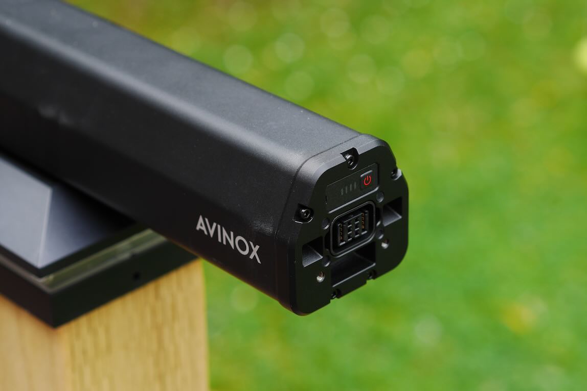 Close-up view of a black device marked 'AVINOX', showcasing its power button and connectors, positioned on a wooden surface with a blurred green background.