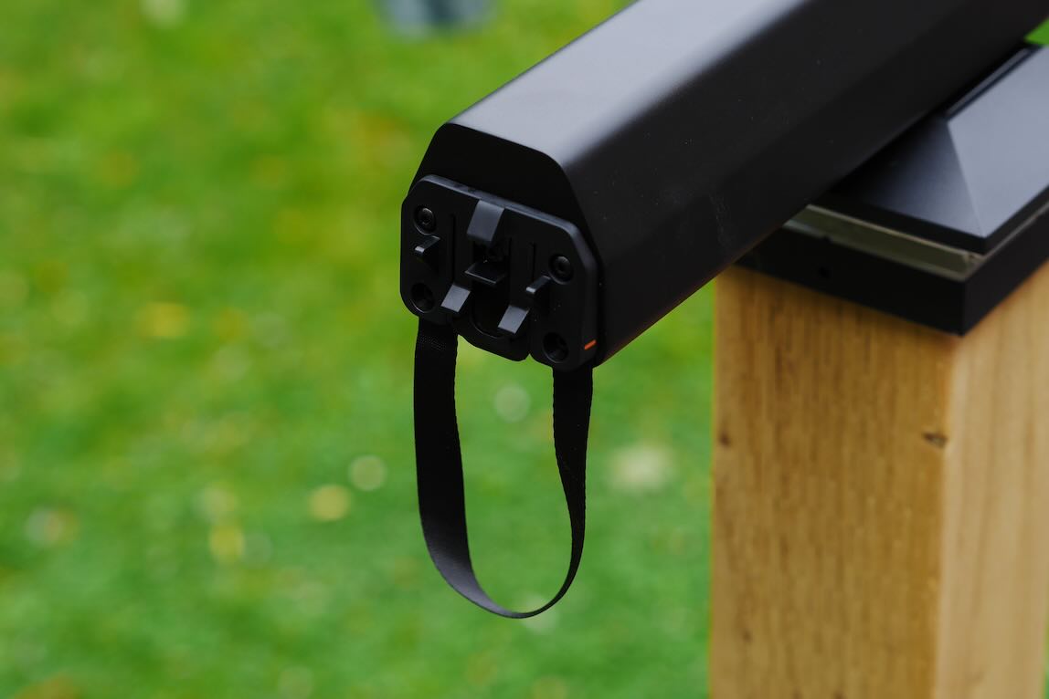 Close-up of a black device with a strap, mounted on a wooden post, set against a green background.