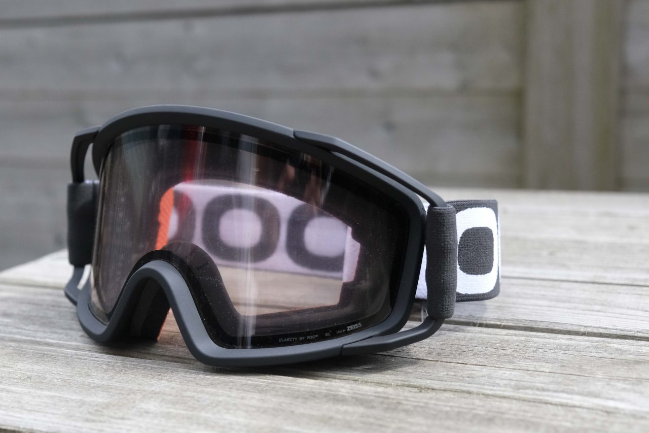 A pair of black ski goggles resting on a wooden surface, featuring tinted lenses and a branded strap.