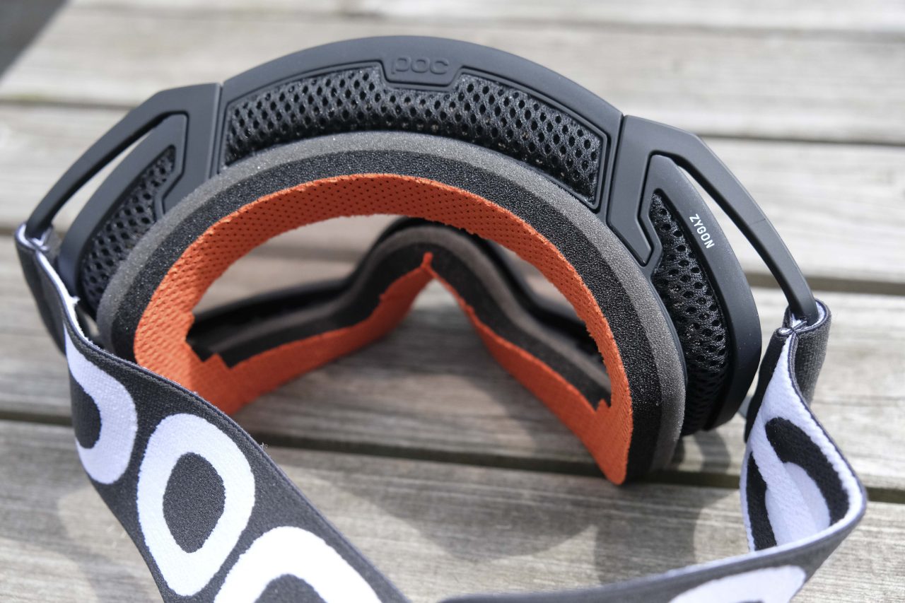 Close-up of a black and orange ski goggle, featuring a padded interior and a branded strap, placed on a wooden surface.