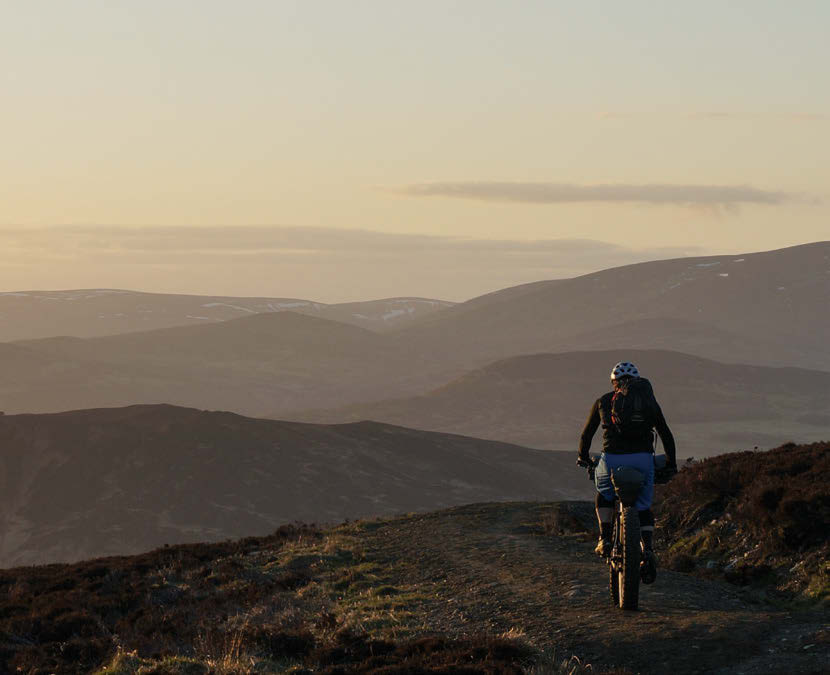 Two bikepackers settle in for dinner at their hilltop camp as dusk falls over the surrounding mountains