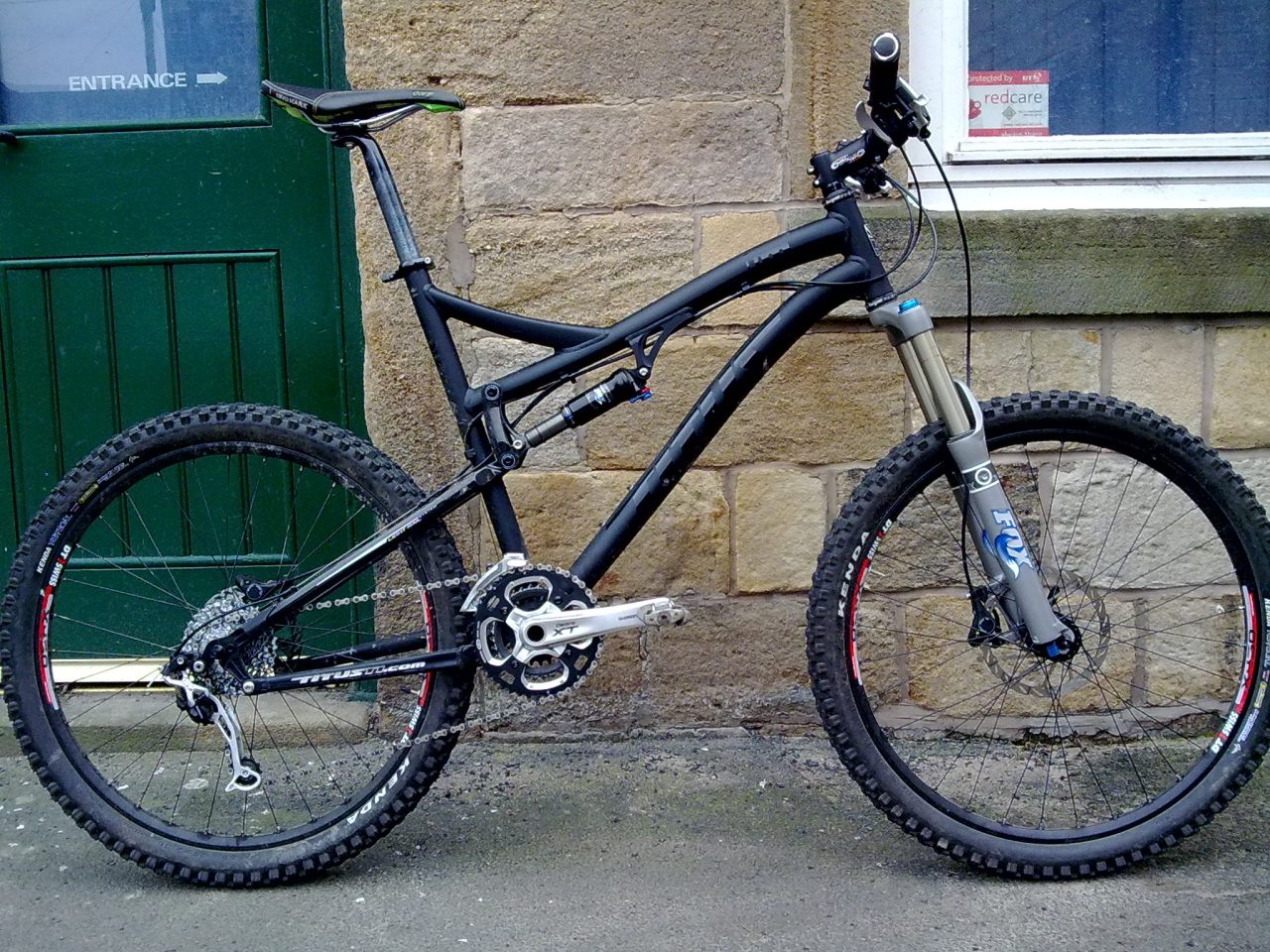 A black mountain bike leaning against a stone wall, with a green door in the background. The bike features thick tyres, a full suspension frame, and a front fork with a shock absorber.