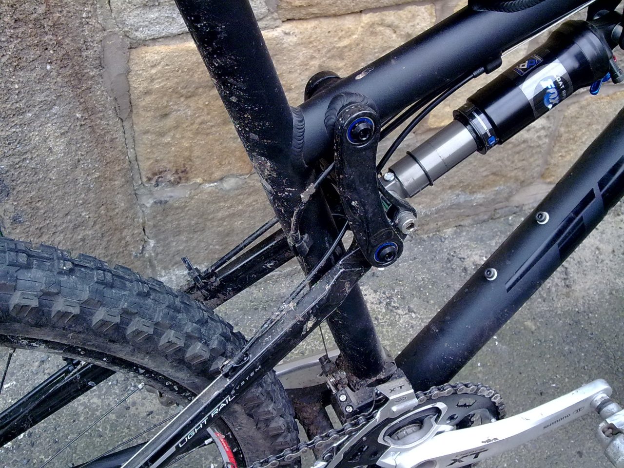 Close-up of a mountain bike frame showing the rear suspension linkage and shock absorber, with visible dirt on the components.