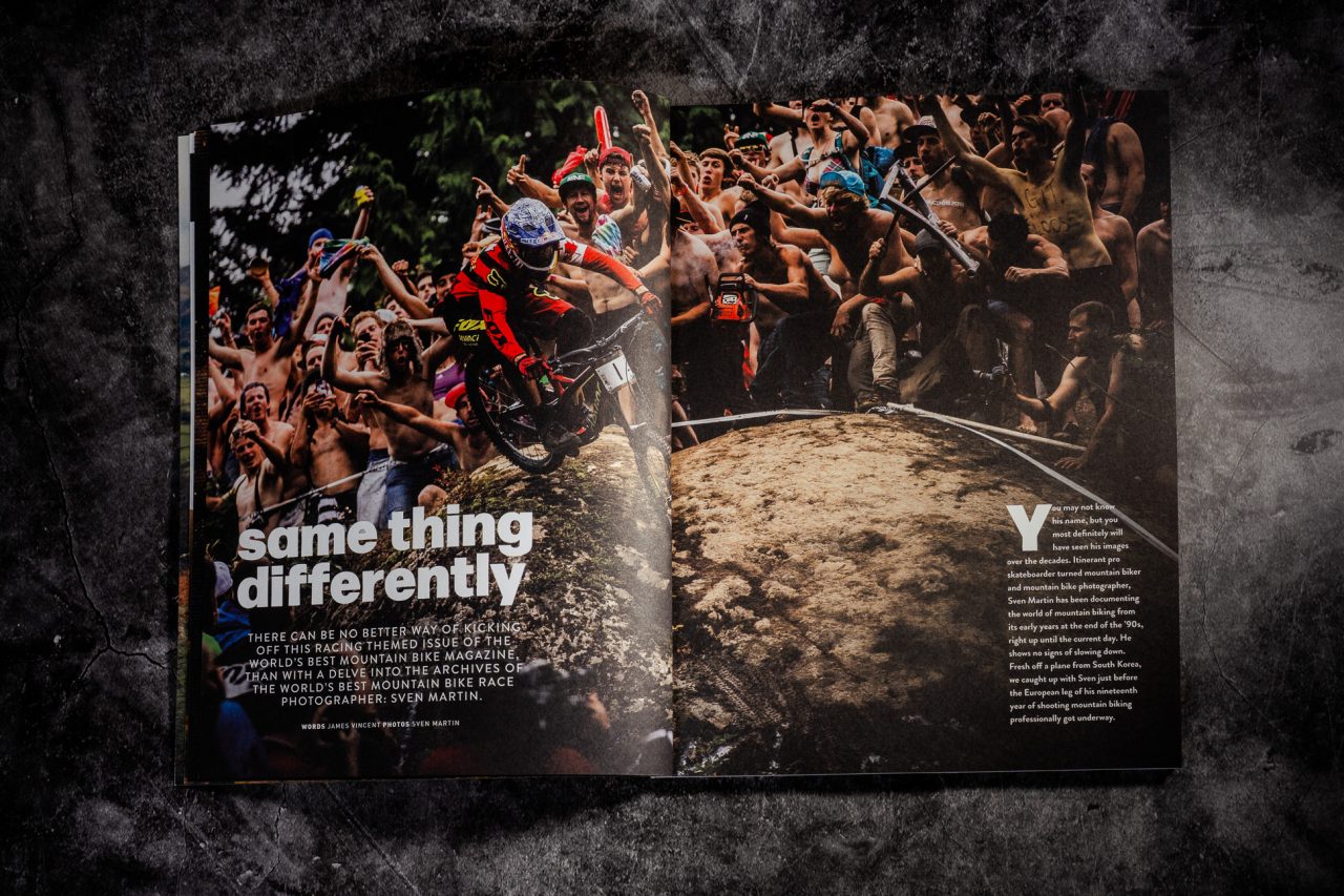 A mountain biker in red gear performs a jump over a rocky terrain, surrounded by an excited crowd of spectators cheering and raising their arms, featured in a magazine spread with text.
