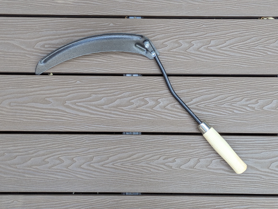 A curved hand scythe tool with a wooden handle lying on a wooden surface.