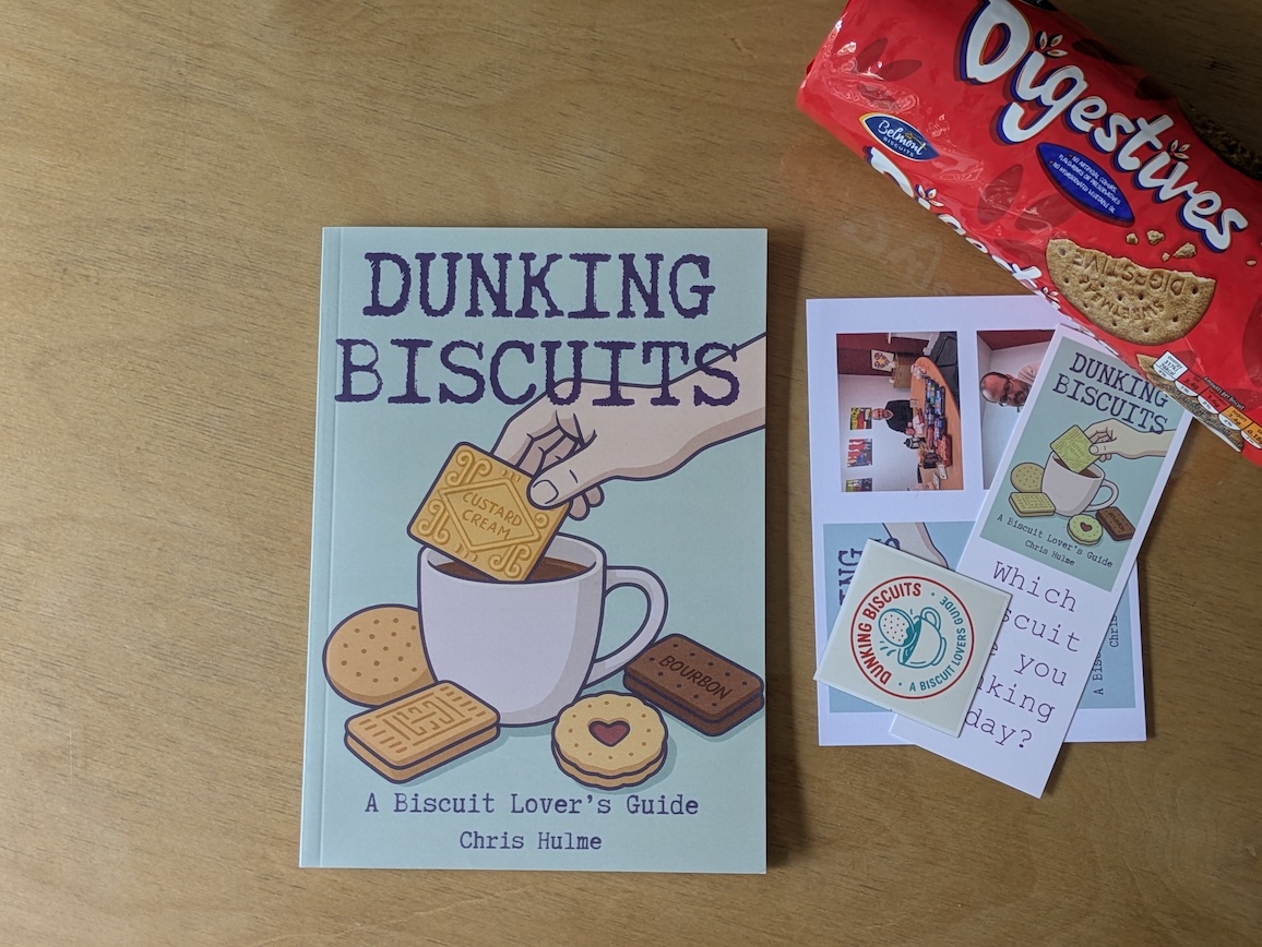 A book titled 'Dunking Biscuits: A Biscuit Lover's Guide' by Chris Hulme, featuring illustrations of biscuits and a hand dunking a Custard Cream into a cup, placed on a wooden surface beside a pack of Digestives and promotional materials.