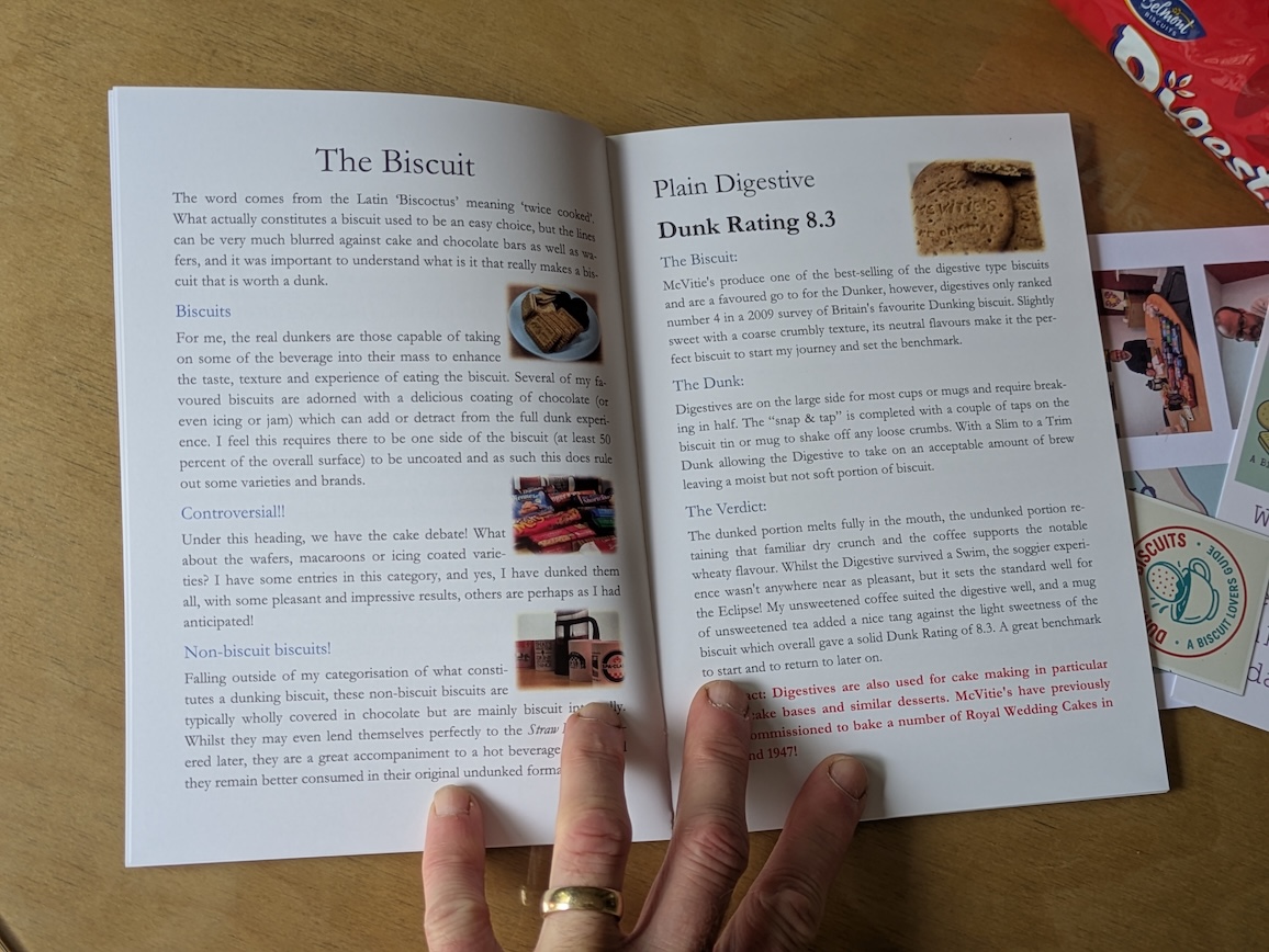 An open booklet titled 'The Biscuit' discussing various aspects of biscuits, featuring a Dunk Rating of 8.3 for Plain Digestive biscuits. It includes text on dunking techniques and flavours, alongside images of biscuits. A hand is visible holding the booklet, with a pack of biscuits partially displayed in the background.