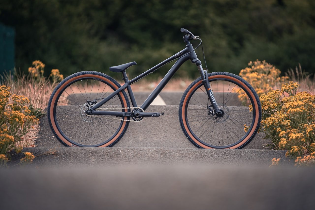 A black mountain bike with brown tyres parked on a concrete surface, surrounded by yellow flowers and green foliage in the background.