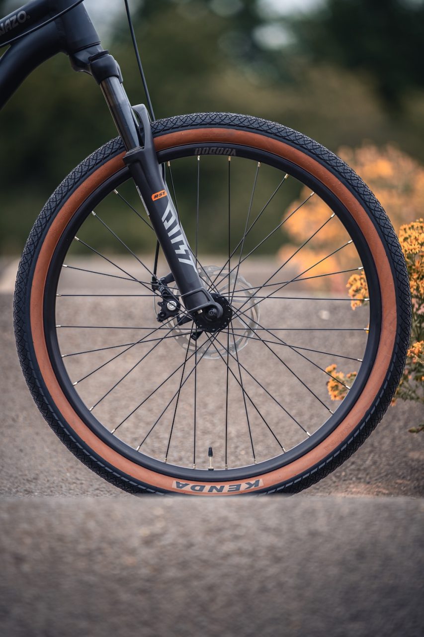 Close-up of a mountain bike wheel with a black and orange tyre, showcasing the spokes and disc brake in a blurred outdoor setting.