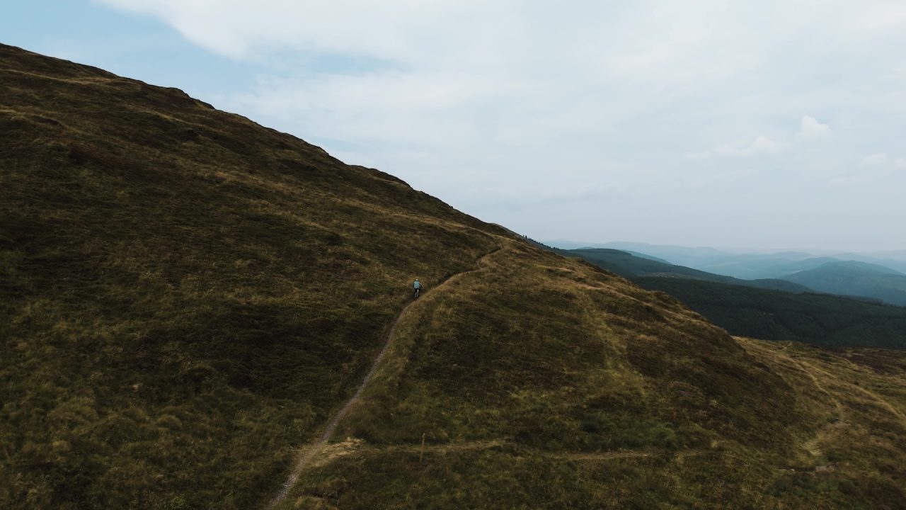 A person walking along a narrow path on a grassy hillside, with rolling hills and a cloudy sky in the background.