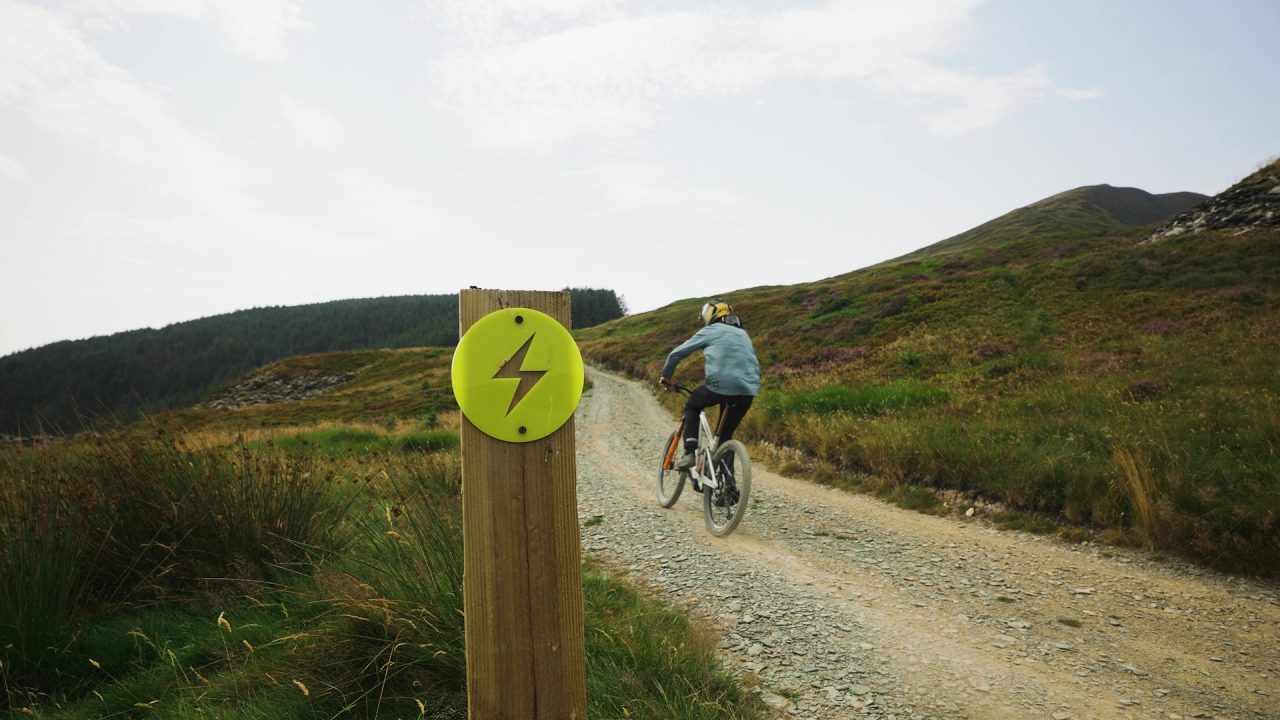 A cyclist riding on a gravel path in a mountainous landscape, with a bright yellow sign featuring a lightning bolt on a wooden post in the foreground.