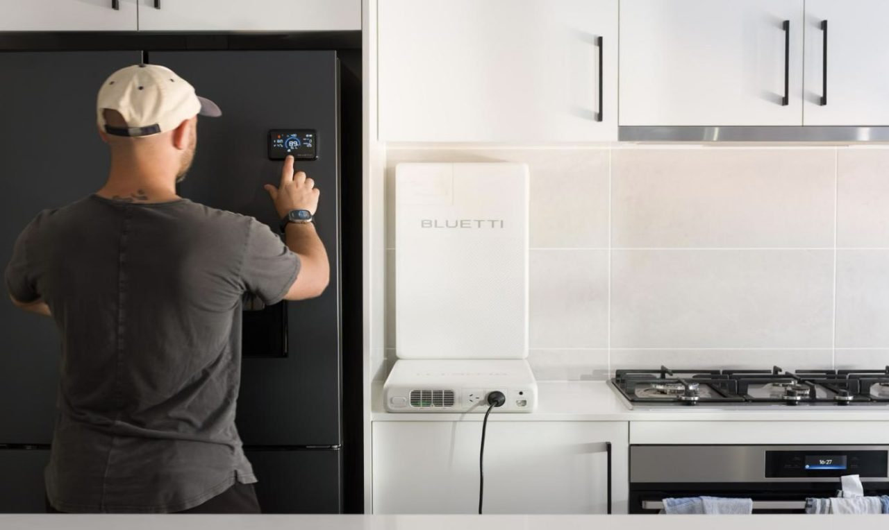 A man adjusting the settings on a modern black refrigerator in a contemporary kitchen, with a white power station and gas stove visible in the background.