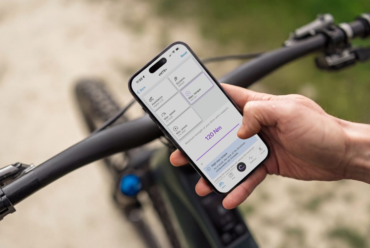 A person holds a smartphone displaying a cycling app while standing next to a mountain bike. The screen shows settings including assistance levels and torque.