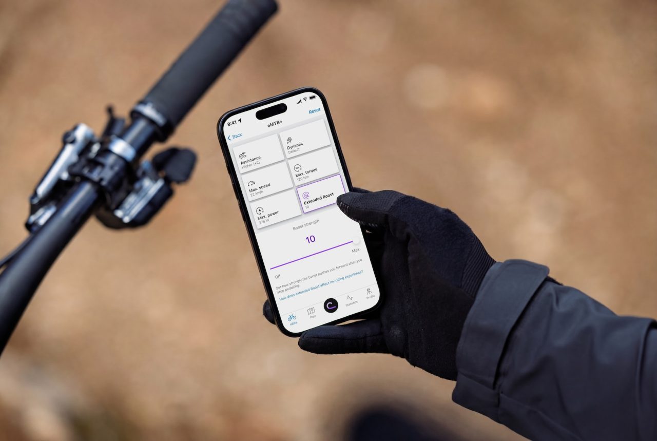 A person in gloves holding a smartphone displaying an eMTB app, with various settings for assistance and power displayed on the screen, next to a bike handlebar.