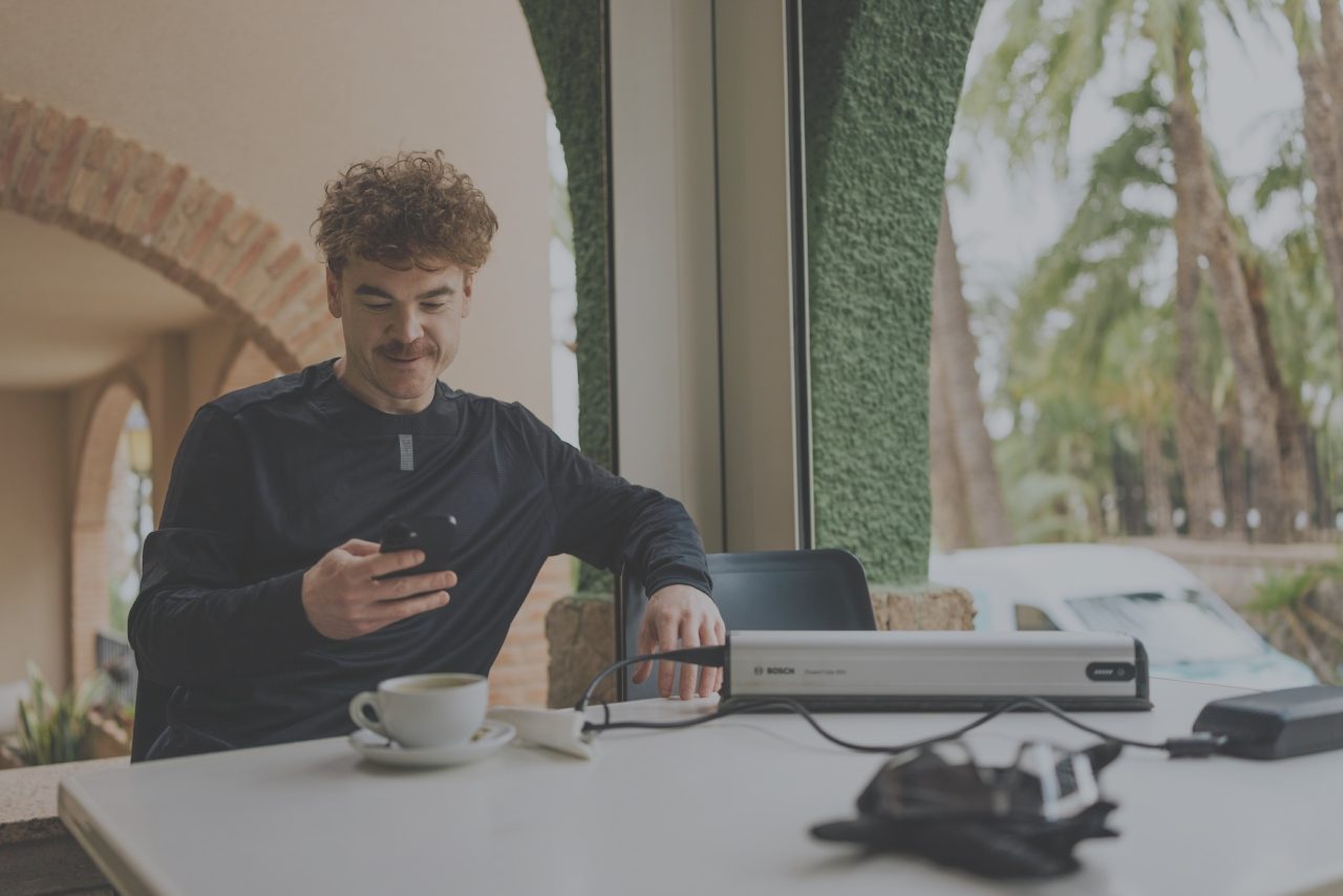 A man with curly hair and a moustache sitting at a table, looking at his smartphone, with a cup of coffee and a scanner visible on the table, and palm trees seen through the window.