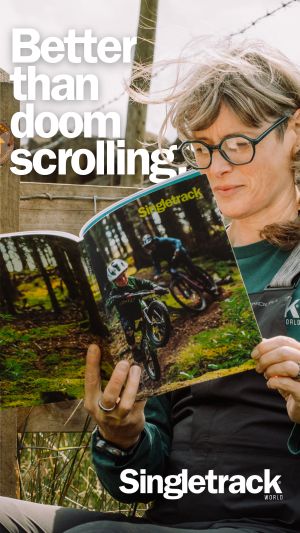 A person reading a magazine titled 'Singletrack' while sitting outdoors, surrounded by nature. The magazine features an image of a cyclist on a mountain bike. The text 'Better than doom scrolling' is prominently displayed.