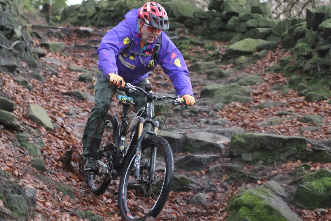 A mountain biker navigating a rocky trail covered in autumn leaves, wearing a purple jacket, helmet, and gloves.