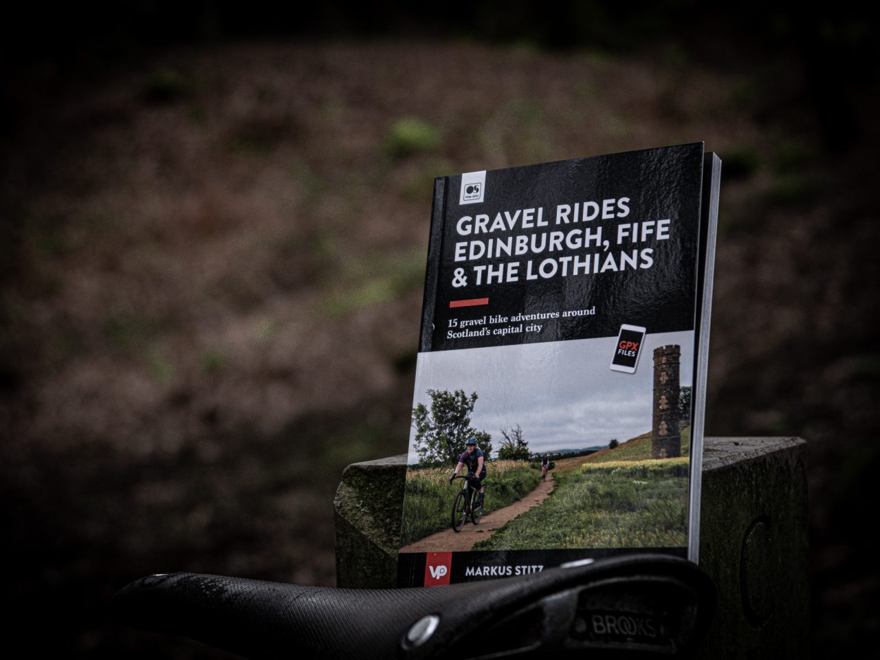 A close-up of a book titled 'Gravel Rides Edinburgh, Fife & The Lothians' resting on a wooden surface, with a bicycle saddle in the foreground. The book features 15 adventure routes around Edinburgh and has a scenic image of cyclists on a gravel path.